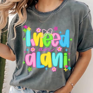 Pode incluir: Camiseta cinza com a frase colorida "I need an alani". O texto &eacute; decorado com flores, borboletas e estrelas. A camiseta tem mangas curtas e gola redonda.