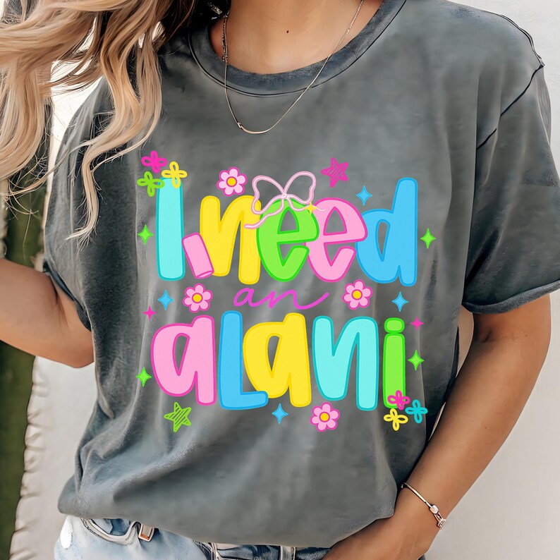 Pode incluir: Camiseta cinza com a frase colorida "I need an alani". O texto &eacute; decorado com flores, borboletas e estrelas. A camiseta tem mangas curtas e gola redonda.