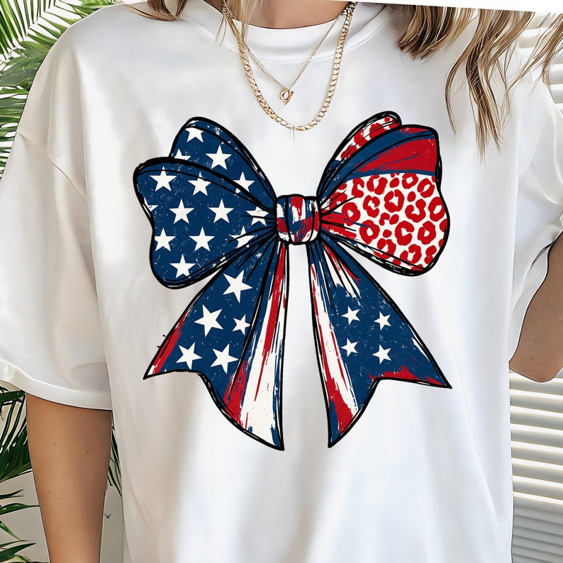 May include: White t-shirt featuring a large bow graphic. The bow design incorporates elements of the American flag with blue star-studded sections, red and white stripes, and a red leopard print pattern.