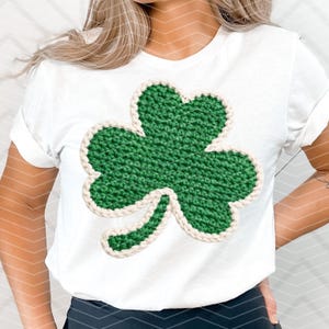 May include: White t-shirt with a large, green crocheted shamrock and a cream-coloured border. The shamrock is centrally placed, giving a festive, handcrafted appearance. Ideal for St. Patrick's Day or general wear.
