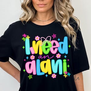 Pode incluir: T-shirt preto com a frase "I need an Alani" em letras coloridas e arredondadas. O design inclui flores, estrelas e um la&ccedil;o. Uma pe&ccedil;a casual para uso di&aacute;rio.