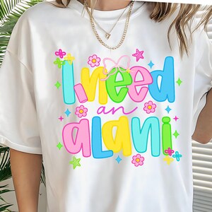 Pode incluir: Camiseta branca com a frase "I need an alani" em letras coloridas. O design inclui flores, estrelas e um la&ccedil;o. &Eacute; uma pe&ccedil;a casual para uso di&aacute;rio.