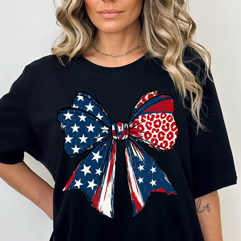 May include: Black t-shirt featuring a large bow design. The bow incorporates elements of the American flag with blue stars, red and white stripes, and a section with a red and white leopard print pattern. The design is centered on the shirt.