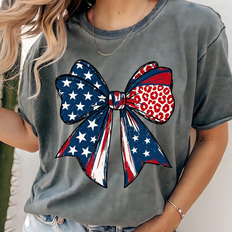 May include: Gray t-shirt featuring a large bow design. The bow incorporates patriotic elements with blue and white stars, red and white stripes, and a section with a red leopard print pattern. The shirt has short sleeves and a crew neck.