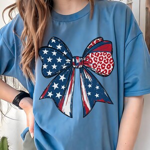 May include: A light blue t-shirt featuring a large bow design. The bow incorporates elements of the American flag with stars, stripes, and a leopard print pattern in red, white, and blue. The shirt is a casual style.