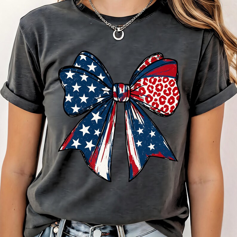 May include: Dark gray t-shirt featuring a patriotic bow design. The bow incorporates the American flag with red and white stripes, white stars on a blue background, and a section with a red and white leopard print.