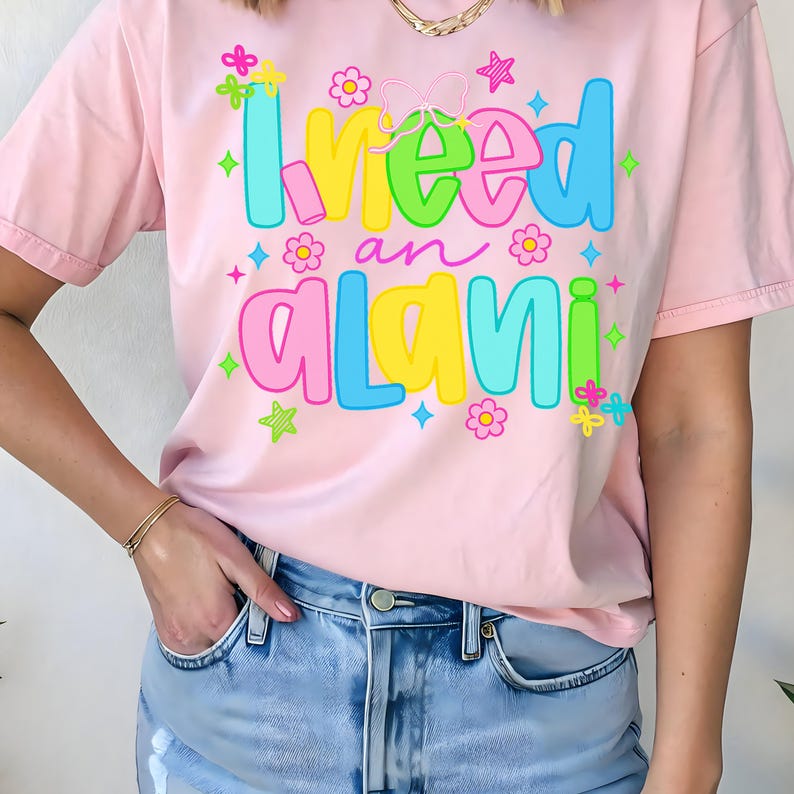 Pode incluir: T-shirt rosa claro com a frase "I need an Alani" em letras coloridas. O design inclui flores, estrelas e um la&ccedil;o. A t-shirt &eacute; feita de um tecido macio, ideal para uso casual.
