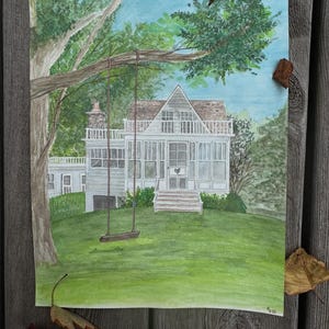 May include: Watercolor painting of a white house with a porch and a swing hanging from a tree branch. The house is on a green lawn, with trees and a blue sky in the background. The painting is on a piece of paper.