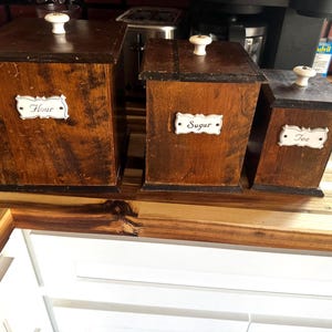 May include: Three wooden kitchen canisters with white labels that read "Flour", "Sugar", and "Tea". The canisters are rectangular with dark brown lids and white knobs. The wood has a distressed finish.