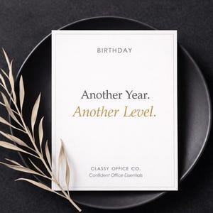 Puede incluir: Una tarjeta de cumpleaños blanca con texto dorado que dice "Another Year. Another Level." está centrada en un plato negro. Una ramita de hojas secas se coloca a la izquierda de la tarjeta. La tarjeta también incluye el texto "Classy Office Co. Confident Office Essentials."
