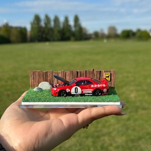 May include: A red model car with a black hood open, sitting on a green grass base with a wooden fence and a sign that says "Beware of Dog". The car has a number "0" on the side and the text "MIDNIGHT MODEL" and "Z-TUNE".