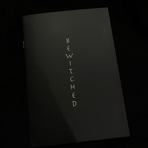 May include: A dark gray notebook with the word "BEWITCHED" written vertically in white letters.