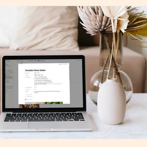 May include: A laptop computer with a white background showing a recipe for Tortellini Pasta Salad on the screen. The laptop is on a white table with a vase of dried flowers and a clear glass mug.