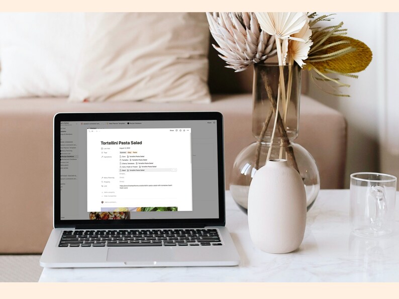 May include: A laptop computer with a white background showing a recipe for Tortellini Pasta Salad on the screen. The laptop is on a white table with a vase of dried flowers and a clear glass mug.