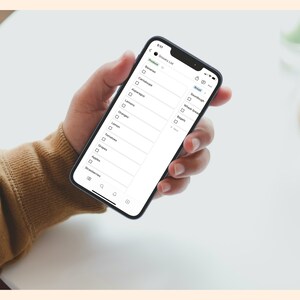 May include: A person's hand holding a smartphone displaying a grocery list app. The list includes items like bananas, asparagus, oranges, and bread.