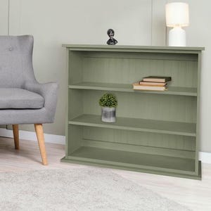 May include: A gray upholstered loveseat with button tufting and wooden legs. A small, round side table with a glass of water and a bowl of fruit sits to the left. A green bookshelf with three shelves and a small lamp are on the right.
