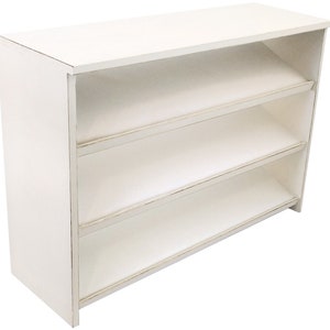 Op de afbeelding: Wit houten rek met drie planken. De planken zijn lichtjes naar voren gekanteld. Het rek heeft een verweerde witte afwerking.