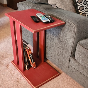 May include: A red wooden end table with a shelf for books. The table has a distressed finish and is sitting next to a gray couch. Three remote controls are on the table top.