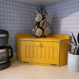 May include: A yellow wooden corner bread box with a drop-down door and a central knob, positioned beneath a black metal mug tree holding six cream-colored ceramic mugs decorated with blue and brown floral patterns and brown rims.