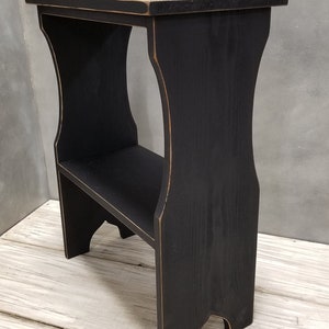 Puede incluir: Un taburete de madera negro con un acabado envejecido. El taburete tiene una parte superior rectangular, un estante inferior y soportes laterales curvos. Los bordes tienen un aspecto m&aacute;s claro y desgastado. El taburete est&aacute; dise&ntilde;ado para usarse como un peque&ntilde;o escal&oacute;n o superficie de exhibici&oacute;n.