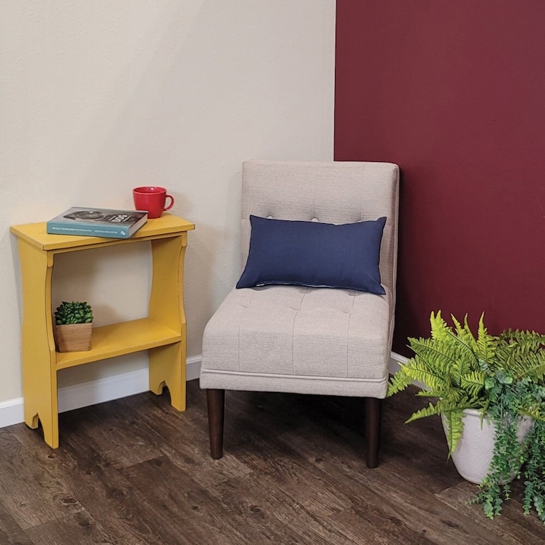 May include: A yellow wooden side table with a lower shelf, a red mug, and a small potted plant. A beige upholstered chair with a navy blue lumbar pillow is next to the table. A fern in a white pot is on the floor.