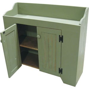 May include: A sage green wooden cabinet featuring a raised back gallery, two doors with round knobs, and black metal hinges. The left door is open, revealing two interior shelves with a natural wood finish. The right door has vertical paneling.