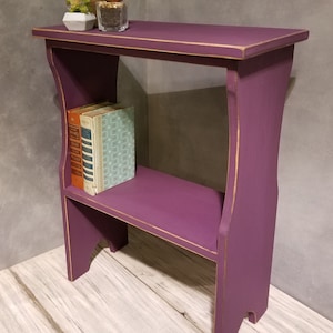 Puede incluir: Peque&ntilde;a mesa auxiliar de madera morada con estante. La mesa tiene ribetes dorados y contiene dos libros y peque&ntilde;as plantas en macetas. La mesa mide aproximadamente 61 cm de alto, 46 cm de ancho y 30 cm de profundidad.