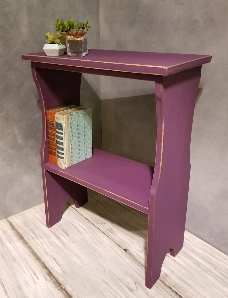 Puede incluir: Peque&ntilde;a mesa auxiliar de madera morada con estante. La mesa tiene ribetes dorados y contiene dos libros y peque&ntilde;as plantas en macetas. La mesa mide aproximadamente 61 cm de alto, 46 cm de ancho y 30 cm de profundidad.