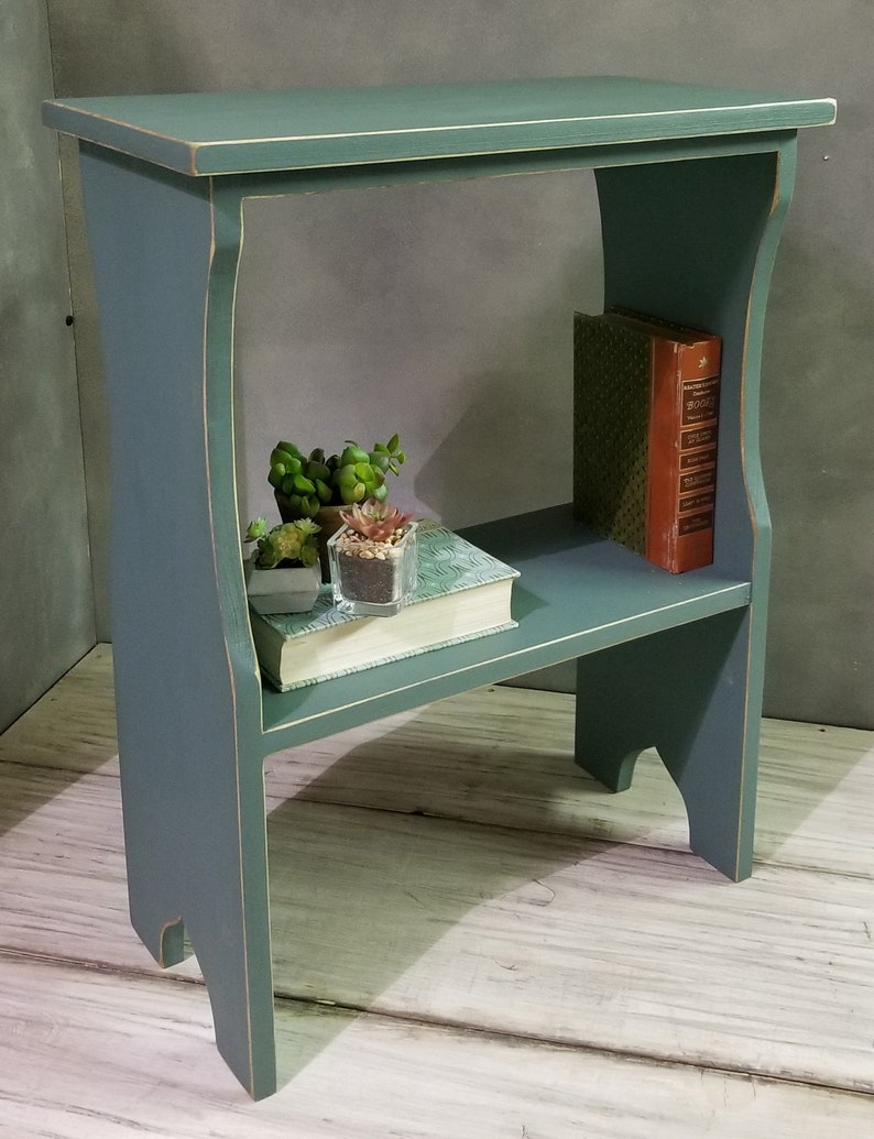 Puede incluir: Una mesa auxiliar de madera de dos niveles, de color verde azulado, con un acabado envejecido. La mesa tiene una superficie superior y un estante inferior, con soportes laterales curvos. El estante contiene un libro y peque&ntilde;as plantas en macetas. La mesa mide 61 cm de alto.