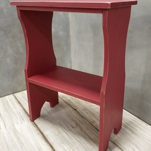 Puede incluir: Una peque&ntilde;a mesa auxiliar de madera roja con un estante. La mesa tiene una parte superior rectangular y un estante inferior, sostenido por patas curvas. El acabado es rojo desgastado, lo que le da un aspecto vintage. La mesa mide aproximadamente 76 cm de alto.