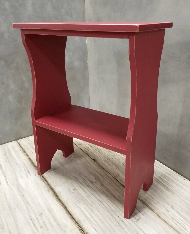 Puede incluir: Una peque&ntilde;a mesa auxiliar de madera roja con un estante. La mesa tiene una parte superior rectangular y un estante inferior, sostenido por patas curvas. El acabado es rojo desgastado, lo que le da un aspecto vintage. La mesa mide aproximadamente 76 cm de alto.
