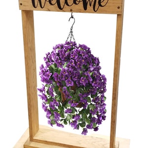 May include: A light brown wooden stand with a rectangular base and a top sign featuring the word Welcome in black script. A hanging basket filled with vibrant purple flowers and green leaves is suspended from the center by a black metal hook.