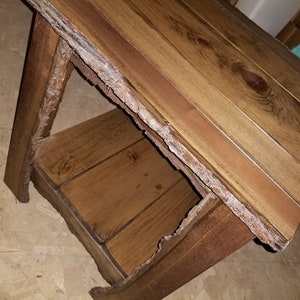 May include: A rustic wooden end table with a natural bark edge and a lower shelf. The table is made of dark brown wood with a distressed finish.