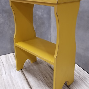 Puede incluir: Peque&ntilde;a mesa auxiliar de madera amarilla con estante. La mesa tiene una parte superior rectangular y un estante inferior, sostenido por patas curvas. El acabado envejecido a&ntilde;ade un aspecto vintage. La mesa mide unos 61 cm de alto.