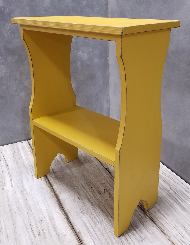 Puede incluir: Peque&ntilde;a mesa auxiliar de madera amarilla con estante. La mesa tiene una parte superior rectangular y un estante inferior, sostenido por patas curvas. El acabado envejecido a&ntilde;ade un aspecto vintage. La mesa mide unos 61 cm de alto.