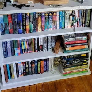 May include: A white bookshelf with three shelves filled with books. The top shelf has a variety of books with different colored spines. The middle shelf has a stack of books with a wooden block on the right side. The bottom shelf has a stack of books on the right side and a variety of books on the left side.