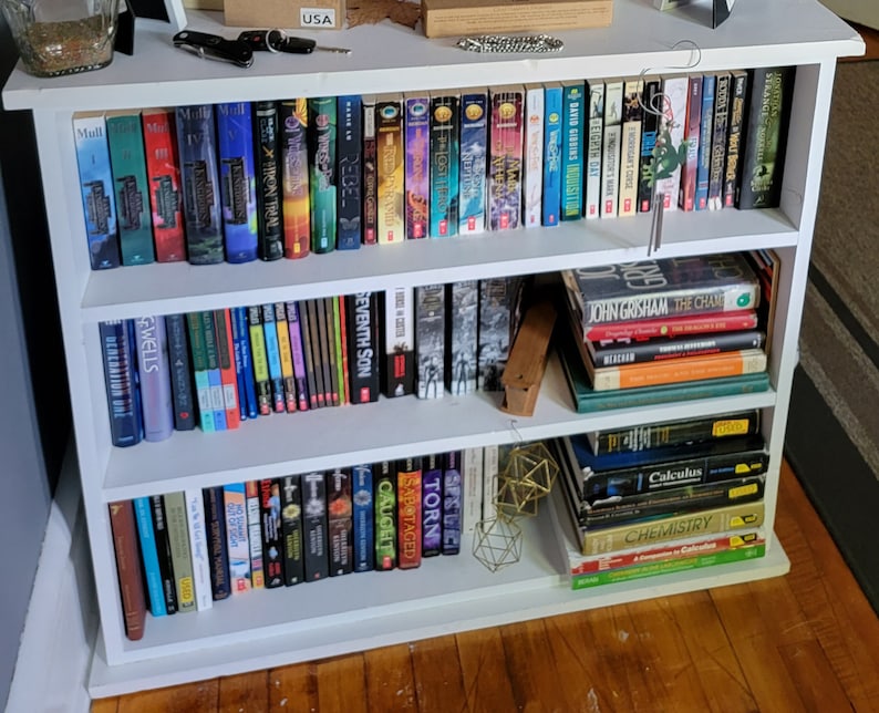 May include: A white bookshelf with three shelves filled with books. The top shelf has a variety of books with different colored spines. The middle shelf has a stack of books with a wooden block on the right side. The bottom shelf has a stack of books on the right side and a variety of books on the left side.
