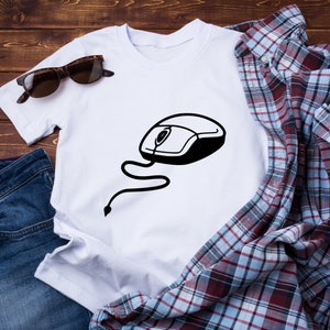 Puede incluir: Una camiseta blanca con un gráfico de ratón de ordenador negro. La camiseta está colocada sobre una superficie de madera con unas gafas de sol, una chaqueta vaquera azul y unos vaqueros azules.