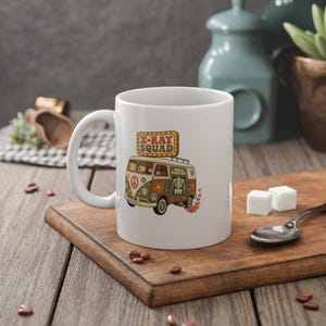 Könnte beinhalten: Ein weißer Keramikbecher mit einer Grafik eines grünen Vintage-Vans, beschriftet mit Mobile X-Ray Unit, verziert mit Blumen und einem Friedenszeichen. Darüber befindet sich ein rechteckiges Schild mit der Aufschrift X-RAY SQUAD in orangefarbenen Buchstaben.