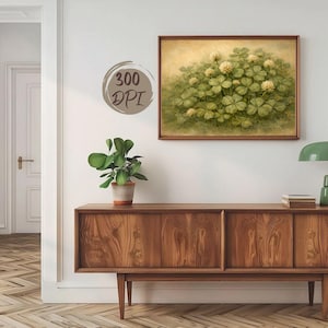 May include: A framed print depicting a cluster of green clover leaves and white flowers. The artwork has a soft, yellow-toned background and is framed in a brown wooden frame. The image is displayed on a white wall above a wooden cabinet.