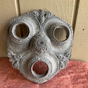 May include: Ornate, gray metal ceiling medallion with three circular openings. The decorative piece features detailed floral and scrollwork patterns. The medallion is likely for lighting fixtures or architectural embellishment.