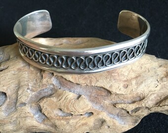 May include: A silver cuff bracelet with a decorative looped design. The bracelet is wide and has a polished finish. The looped design is a repeating pattern around the center of the bracelet. The bracelet is displayed on a piece of driftwood.