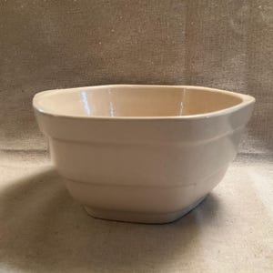 May include: A large, off-white ceramic mixing bowl with a rounded, octagonal shape. The bowl has a smooth, glazed finish and is slightly rippled around the sides.
