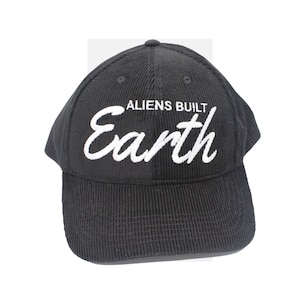 Op de afbeelding: Zwarte corduroy baseballpet met witte geborduurde tekst die "ALIENS BUILT Earth" in een cursief schrifttype luidt.