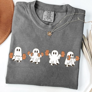 May include: A gray Comfort Colors t-shirt featuring a graphic of four white ghosts lifting orange barbells. The shirt has a crew neck and short sleeves. A silver necklace with a pendant is also visible.