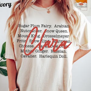 May include: Ivory-colored t-shirt with a list of names in black text, including "Sugar Plum Fairy" and "Nutcracker." The word "Clara" is written in red cursive across the center of the shirt. The word "Ivory" is in the top left corner.