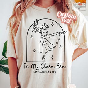 Könnte beinhalten: Ein elfenbeinfarbenes T-Shirt mit einer schwarzen Strichzeichnung einer Ballerina, die einen Nussknacker in einem bogenförmigen Rahmen mit Sternen hält. Unter der Grafik steht der Text In My Clara Era und NUTCRACKER 2026. Ein Etikett zeigt Custom Text.