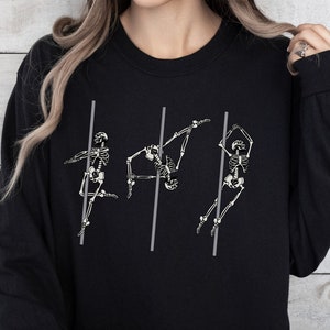 May include: Black sweatshirt featuring a graphic of three skeletons performing a pole dance. The skeletons are white and the poles are grey.