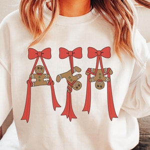 Może przedstawiać: Biała bluza z uroczym wzorem przedstawiającym trzech piernikowych ludzików zawieszonych na czerwonych kokardach i wstążkach. Słowo "White" jest wydrukowane ozdobną czcionką na dole. Przytulny element odzieży świątecznej.