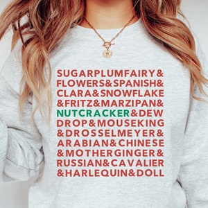 May include: Ash-colored sweatshirt with a list of Nutcracker characters in red and green text. The text includes "SUGARPLUMFAIRY", "NUTCRACKER", and "HARLEQUIN". The sweatshirt is paired with blue jeans.